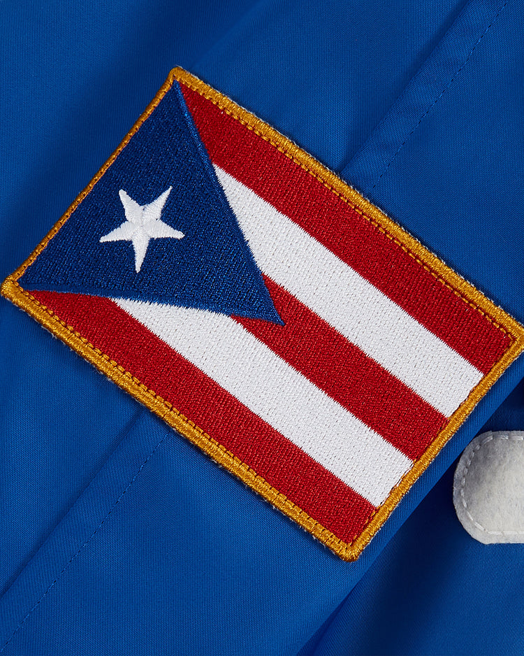 The Ebbets Field Santurce Cangrejeros Cotton Lined Satin Varsity Jacket features a Puerto Rican flag patch with red and white stripes, a blue triangle, and a white star, sewn onto blue fabric in vintage varsity style.