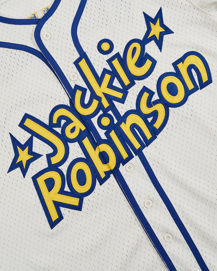 The Ebbets Field Jackie Robinson Comic Book Hero Button-Up Mesh Jersey is white with blue buttons and trim, featuring “Jackie Robinson” in bold yellow and blue letters with star accents across the front.