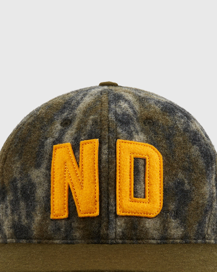 A close-up of the Ebbets Field Mossy Oak x 1966 University of Notre Dame Ballcap shows large yellow-orange ND letters on camouflage, inspired by the classic 1966 Notre Dame design. The background is plain light gray.