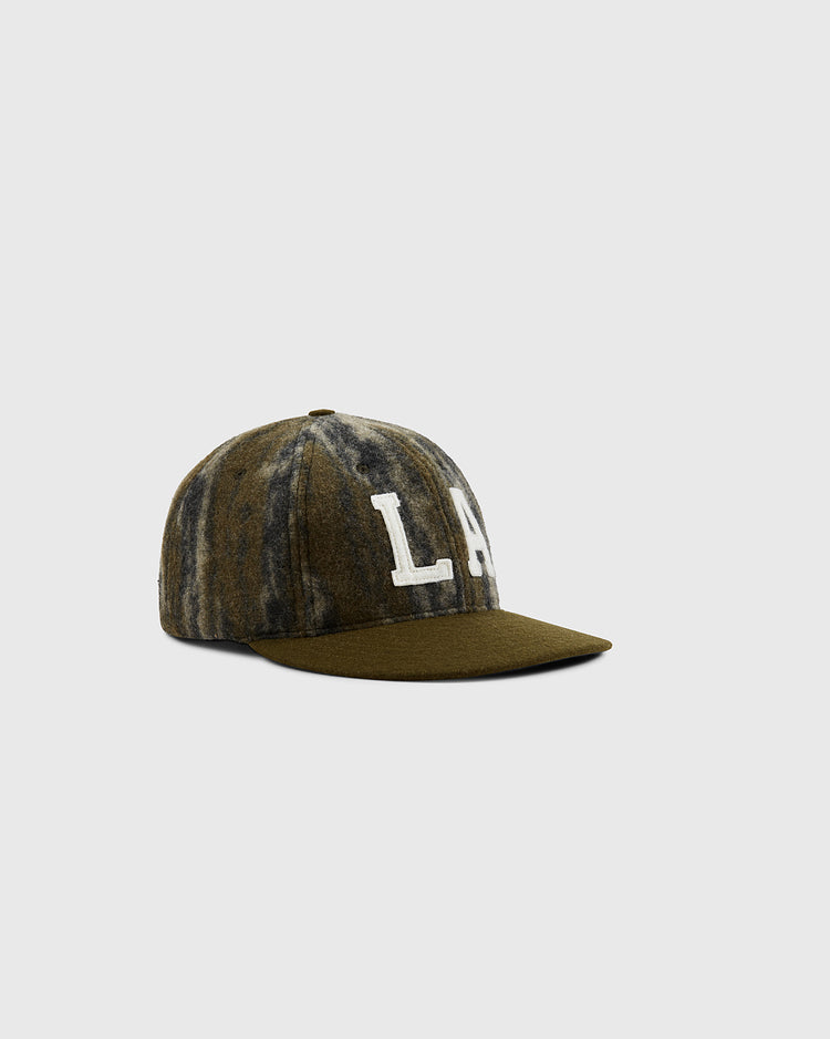 The Ebbets Field Mossy Oak x Ebbets Field 1949 Los Angeles Angels (PCL) Ballcap features a green and gray tie-dye design, flat brim, large white LA embroidery on the front, and is displayed on a plain light gray background.