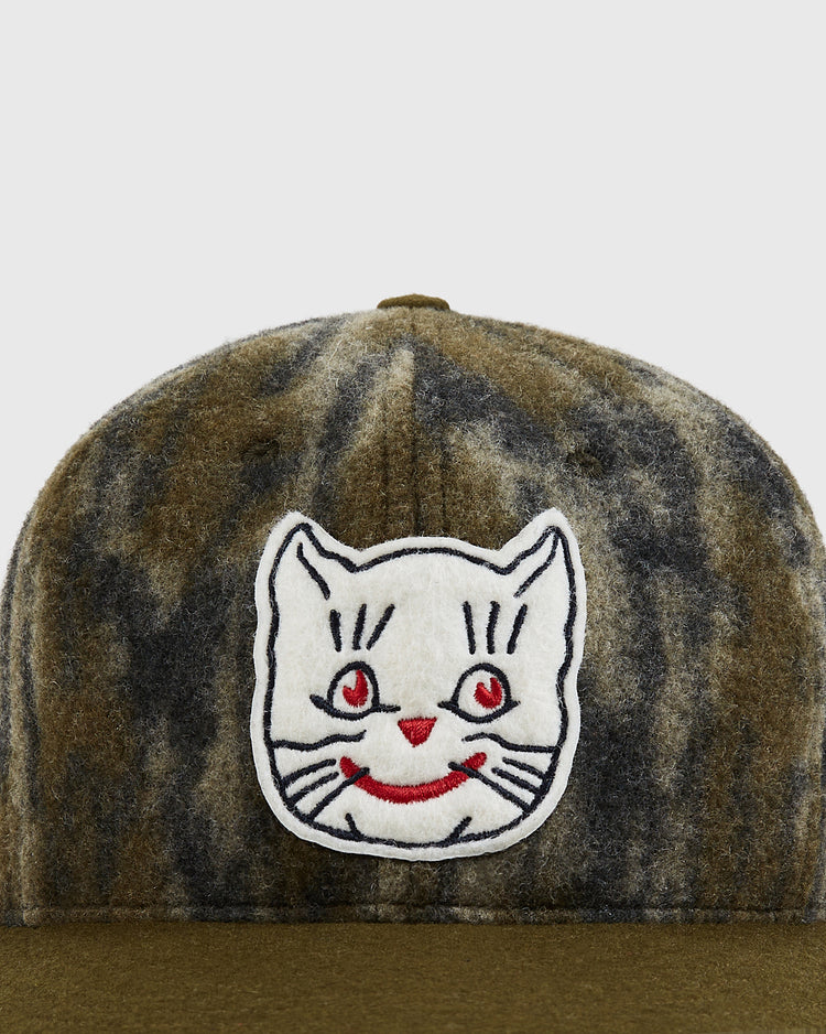 A close-up of the Mossy Oak x Ebbets Field 1961 Kansas City Katz Ballcap by Ebbets Field, featuring a mottled olive and gray pattern and a white patch embroidered with a smiling cat’s face with red eyes and mouth on the front.