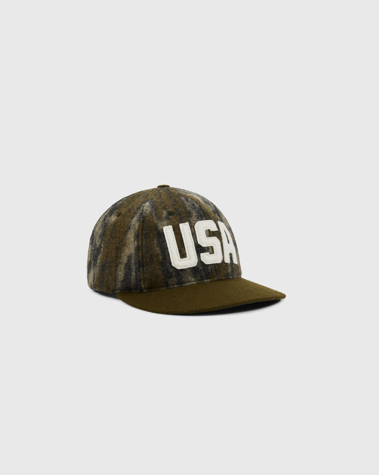 The Ebbets Field Mossy Oak x 1956 USA National Team Ballcap features Mossy Oak Bottomland camo, an olive brim, and bold white USA letters on the front, shown against a plain light gray background.