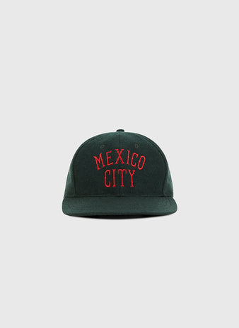 The Ebbets Field Mexico City City Series Wool Ballcap is a dark green hat featuring MEXICO CITY embroidered in red capital letters on the front, shown against a plain light gray background.