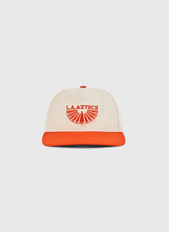 The Ebbets Field Los Angeles Aztecs A-Frame Cotton Twill Ballcap features a beige structured snapback, orange brim, and “L.A. AZTECS” with a sunburst graphic—paying tribute to classic North American Soccer League style.