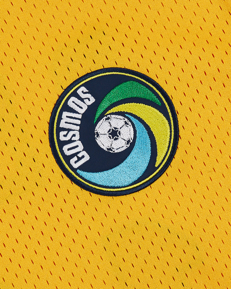 Yellow mesh jersey from Ebbets Field with a circular soccer ball patch surrounded by blue, green, and yellow swirls. “COSMOS” is printed on the side, inspired by the iconic New York Cosmos and classic Pelé jersey.