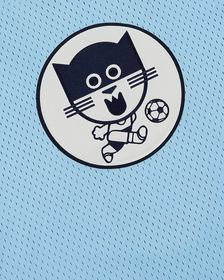 The Ebbets Field Chicago Cats Mesh Soccer Jersey features a circular patch of a smiling cartoon cat kicking a soccer ball, reflecting the playful spirit of the team. The patch stands out on the light blue mesh fabric.
