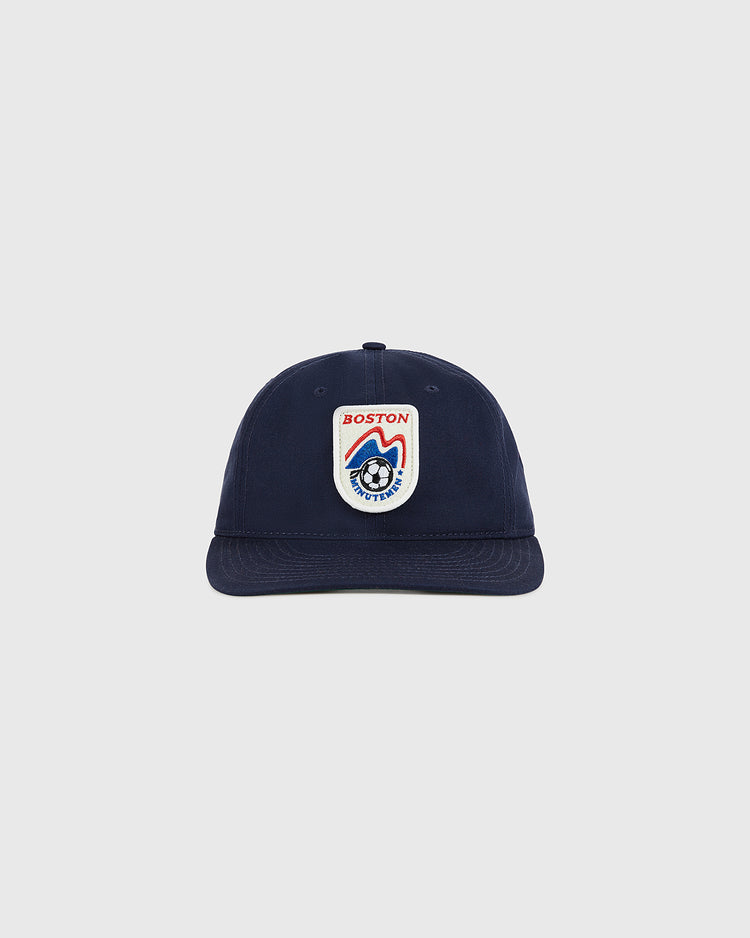The Ebbets Field Boston Minutemen Cotton Twill Ballcap is navy blue with a front patch showing a red and white shield, 