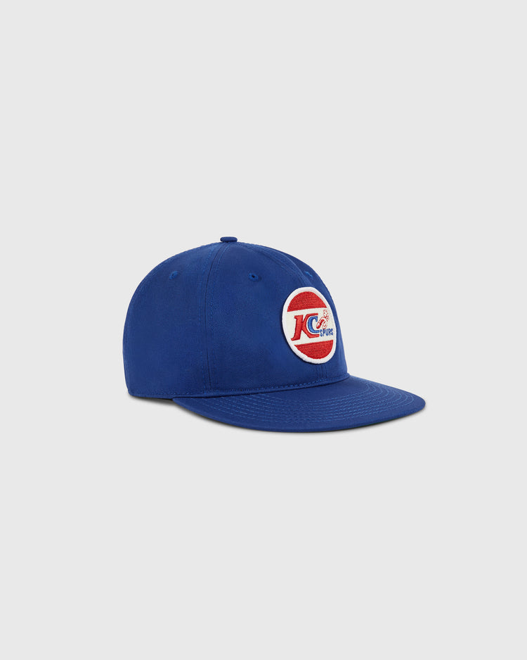 The Ebbets Field Kansas City Spurs Cotton Twill Ballcap is blue with a circular patch featuring a red “C,” white “KC,” and flying baseball, honoring the team’s NASL championship. Shown on a light grey background.