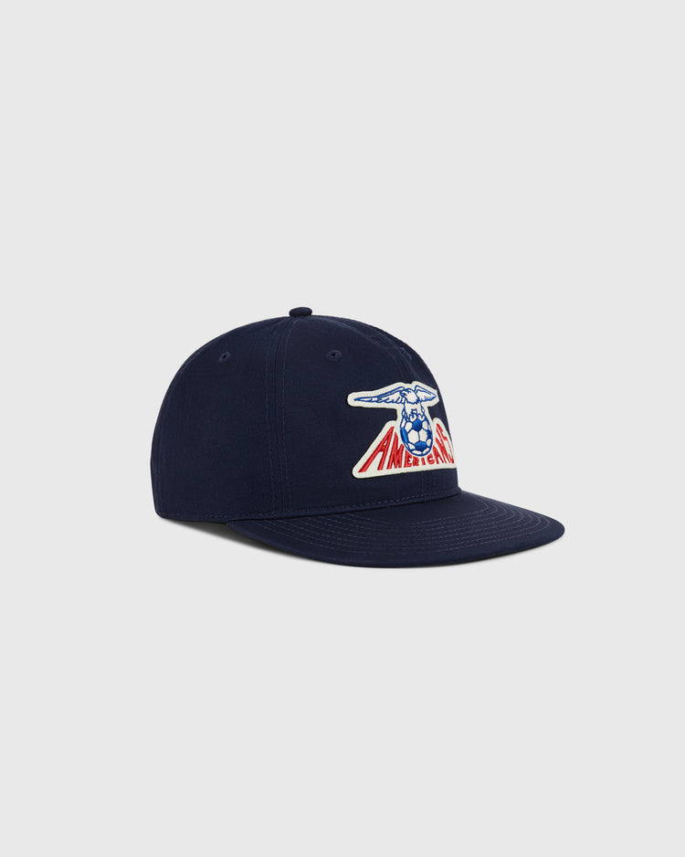 The New Jersey Americans Cotton Twill Ballcap by Ebbets Field features a navy blue base, curved brim, button top, and an embroidered patch of a winged soccer ball above “AMERICANO” in red and blue, inspired by classic ASL championship hats.