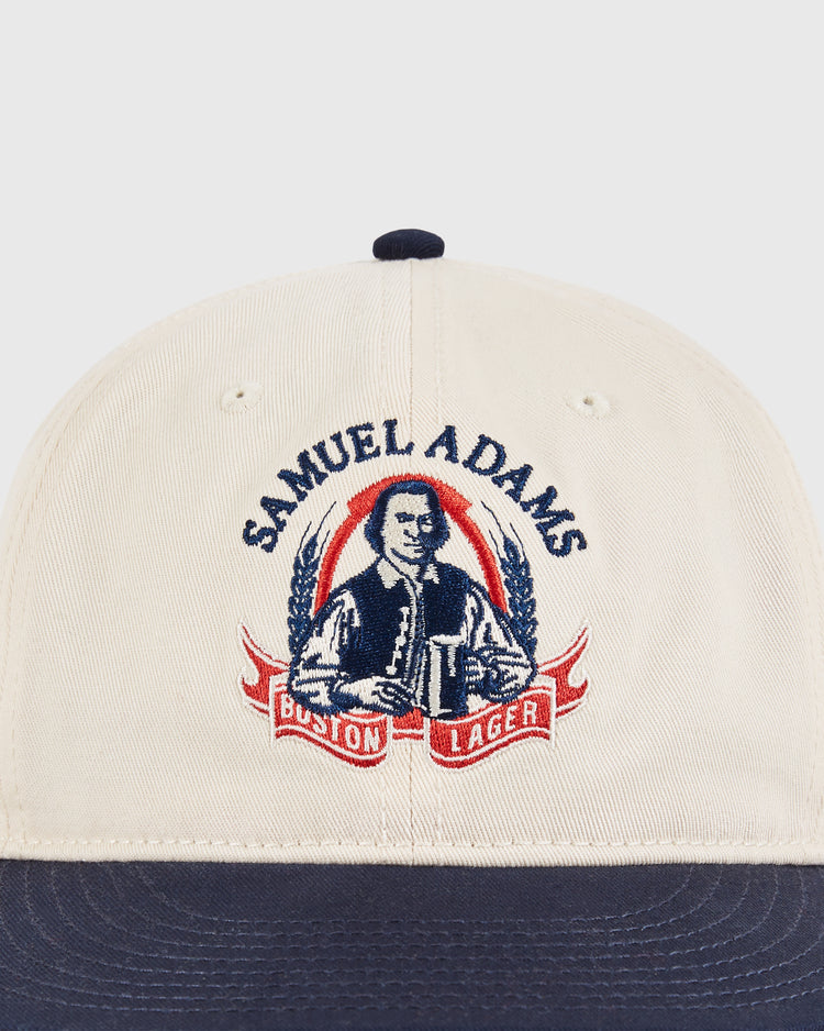 The Ebbets Field Sam Adams Crest Cotton Twill Ballcap features a beige and navy design with a vintage crest logo of a man holding a beer mug, “Samuel Adams” above, and “Boston Lager” in a red ribbon below.