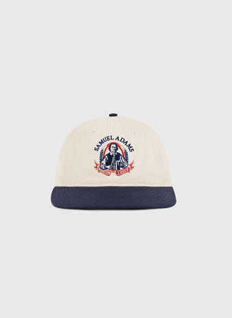 The Sam Adams Crest Cotton Twill Ballcap by Ebbets Field is beige with a navy brim, featuring a vintage crest and an illustration of a man holding beer mugs on the front.