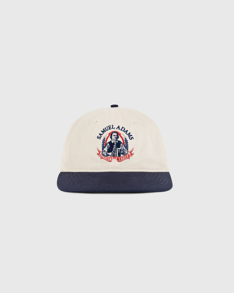 The Sam Adams Crest Cotton Twill Ballcap by Ebbets Field is beige with a navy brim, featuring a vintage crest and an illustration of a man holding beer mugs on the front.