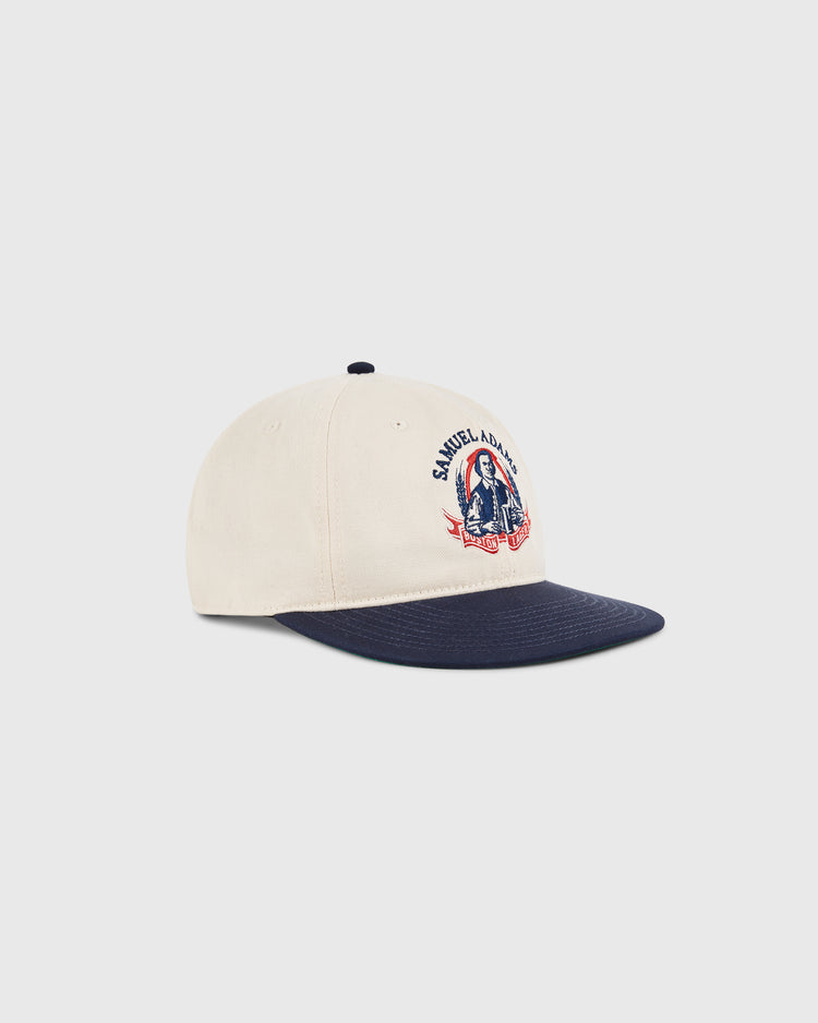 The Ebbets Field Sam Adams Crest Cotton Twill Ballcap features a beige crown, navy blue brim, vintage Samuel Adams crest, and an illustration of a man holding a beer mug on the front.