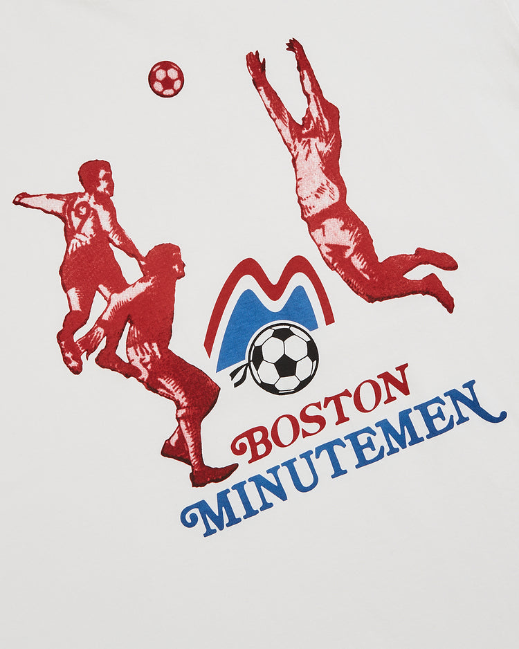 The Ebbets Field Boston Minutemen Short Sleeve Graphic T-Shirt features a red illustration of three soccer players, a ball, and the team name—ideal for any vintage soccer shirt collection or fans of the Boston Minutemen.