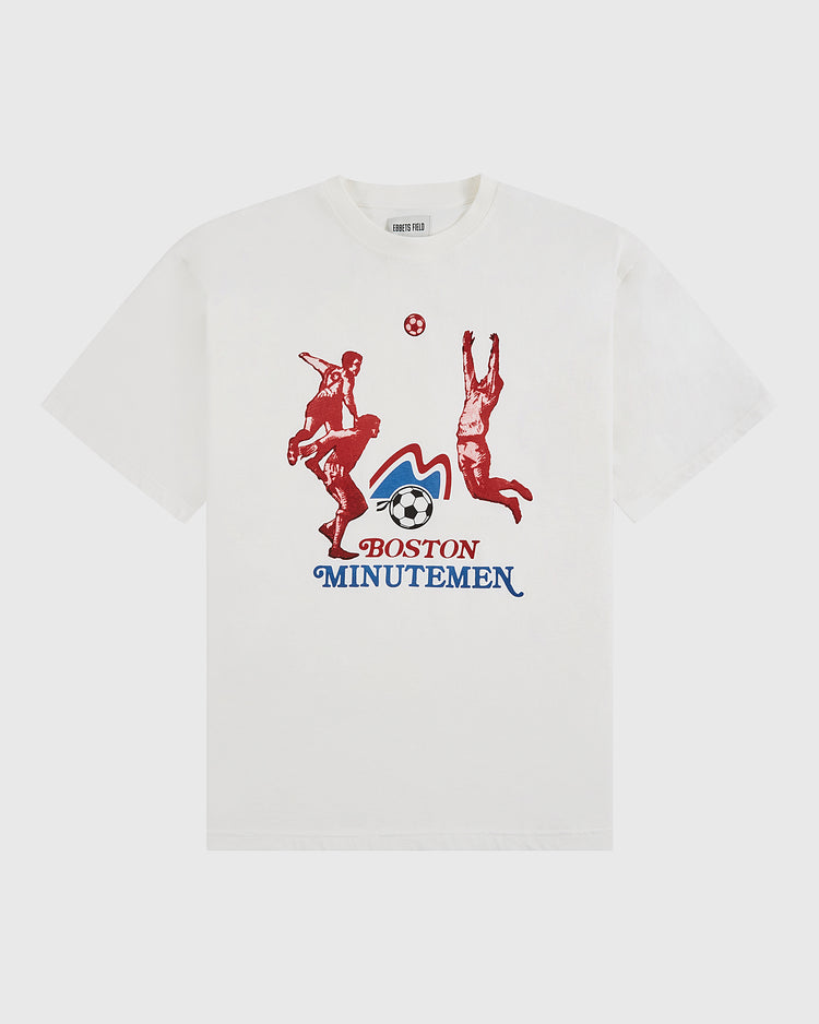 The Ebbets Field Boston Minutemen Short Sleeve Graphic T-Shirt features red soccer player silhouettes, a blue and red soccer ball logo, and bold team text—ideal for fans of vintage soccer shirts.