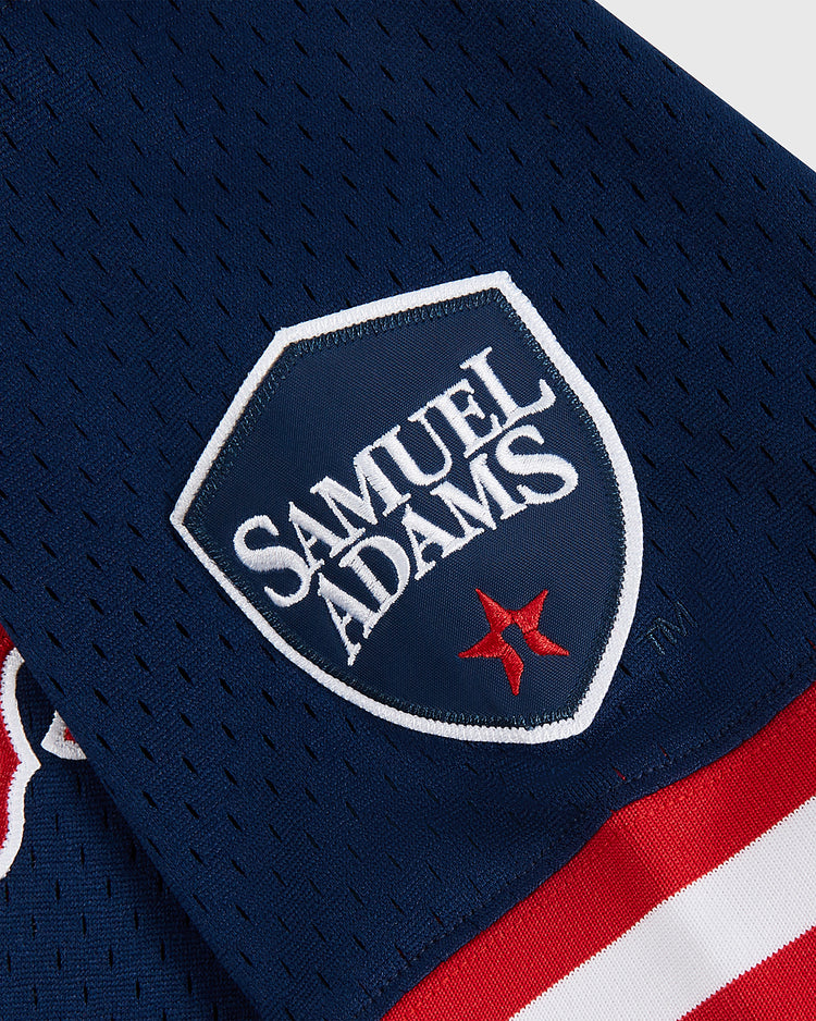 Close-up of the Sam Adams Mesh V-Neck Jersey by Ebbets Field in navy blue, featuring a shield patch with Samuel Adams in white, a red star beneath, and white/red stripe borders.
