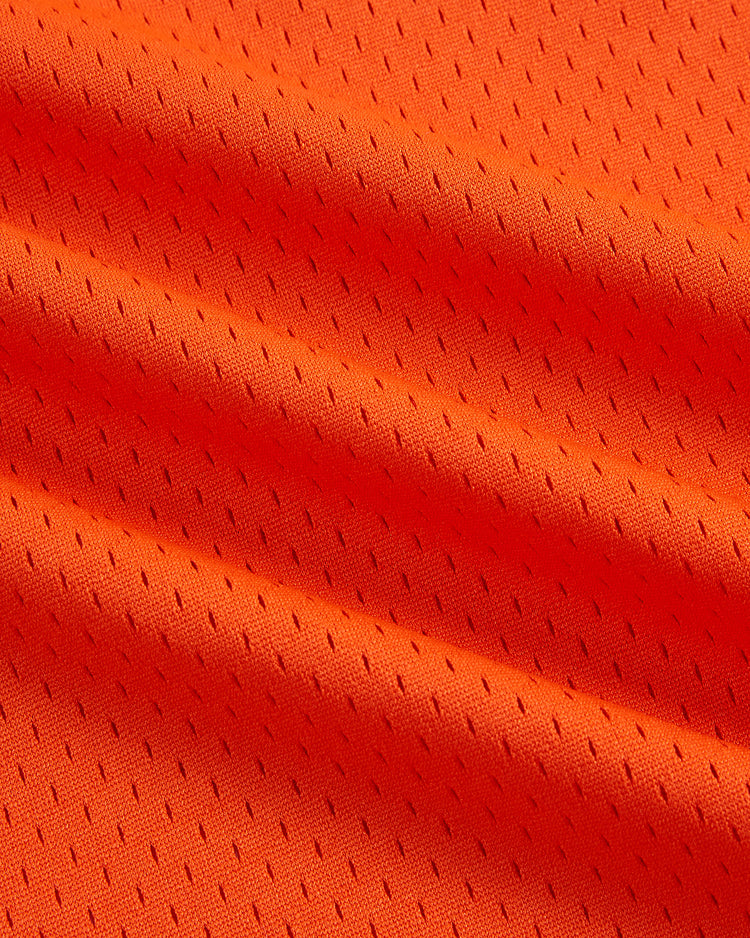 Close-up of vibrant orange mesh fabric from the Ebbets Field Cal Ripken Jr. Charlotte Orioles Mesh V-Neck Jersey, featuring small, evenly spaced perforations and visible folds that create dynamic shadows and texture across the surface—a nod to the iconic jersey style worn by Ripken during his minor league days.