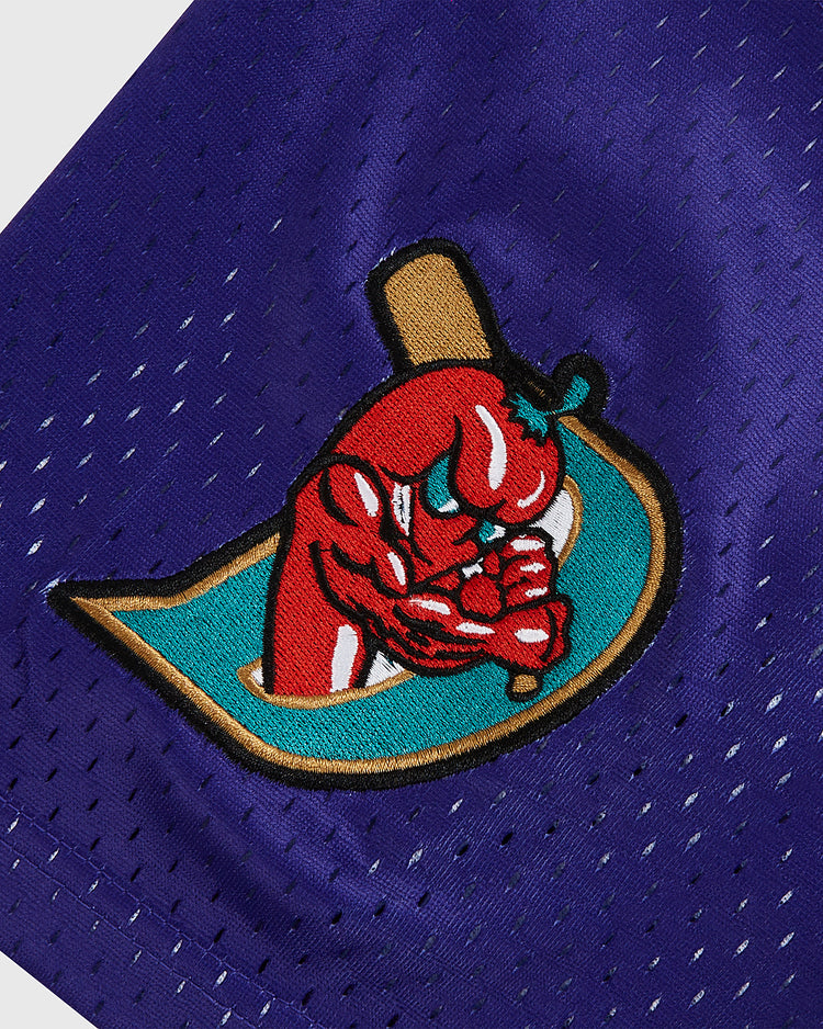 A close-up of a blue fabric patch shows a muscular red chili pepper with crossed arms, gripping a baseball bat and wearing gloves, set against a turquoise background—capturing the bold spirit found on the Ebbets Field Randy Johnson El Paso Diablos Button Up Mesh Jersey.