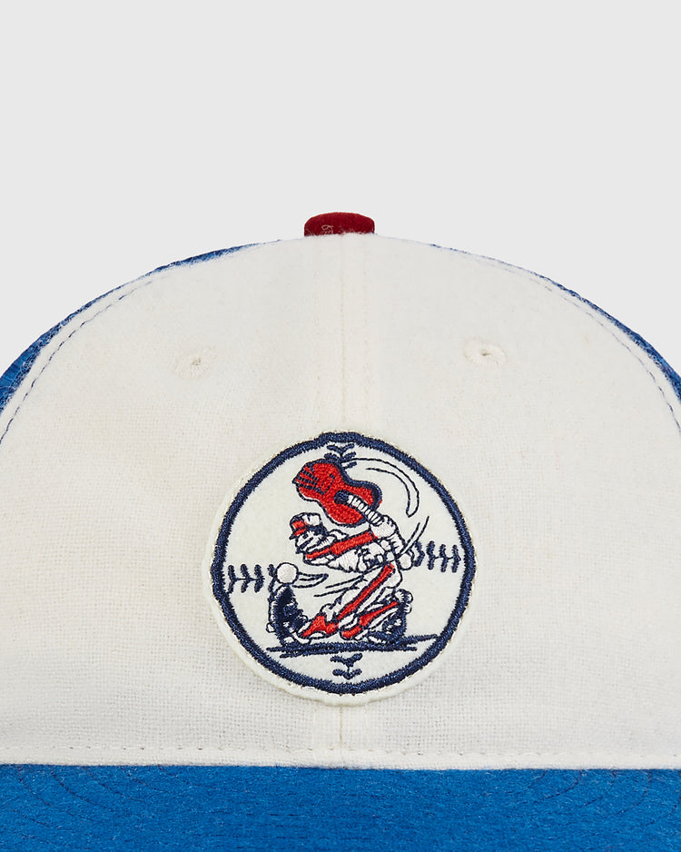 A close-up of the Ebbets Field Nashville Sounds Mesh Trucker Ballcap, featuring a white and blue design with a circular logo depicting a red bird swinging a bat over stitched lines that resemble a baseball.