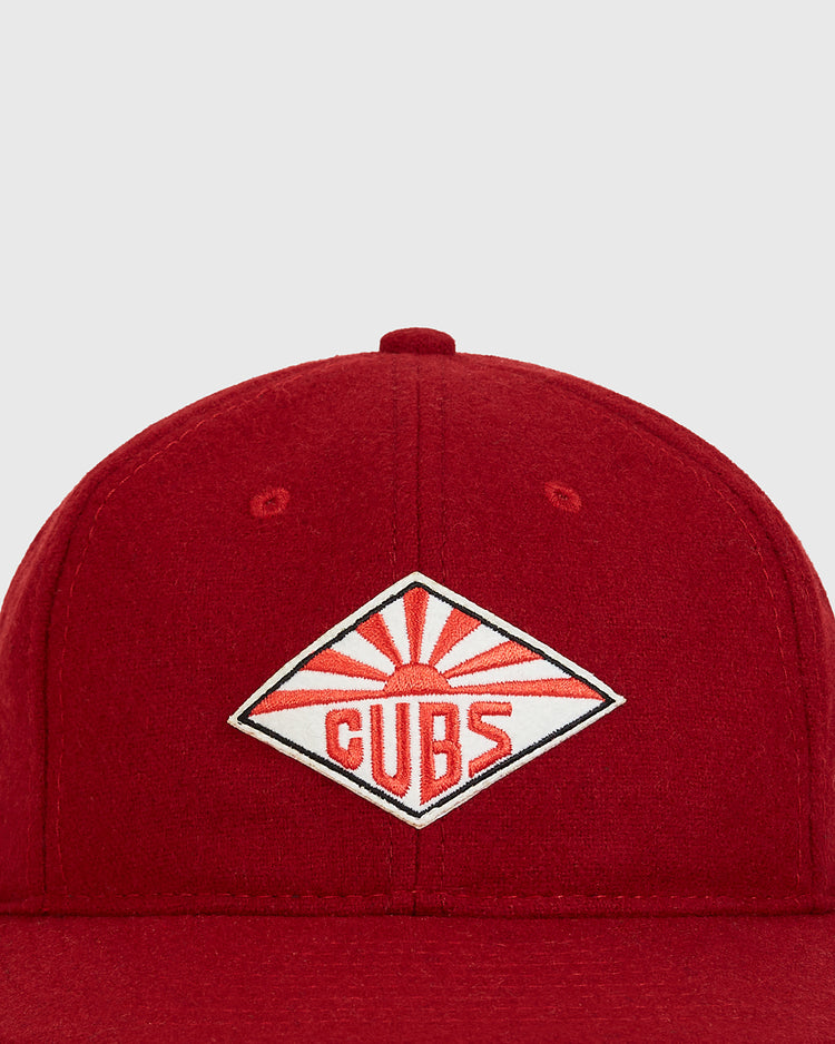 The Ebbets Field Taiyo Cubs 1928 Authentic Wool Ballcap is a red cap featuring a diamond-shaped patch with a stylized red sunburst and the word “CUBS” in red letters at the center.