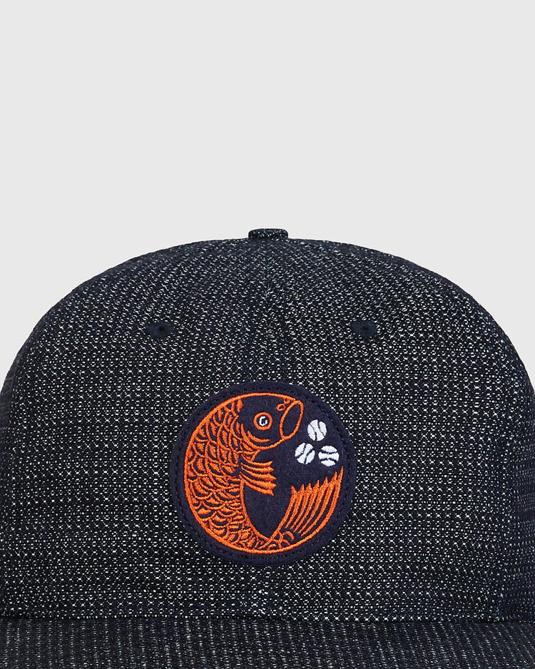 A close-up of the Ebbets Field Hiroshima Carp Japanese Indigo Cotton Dobby Ballcap showcases its premium Japanese cotton construction, accented by an orange embroidered fish with white flowers inside a circular patch on the front—a subtle nod to the iconic Hiroshima Carp.