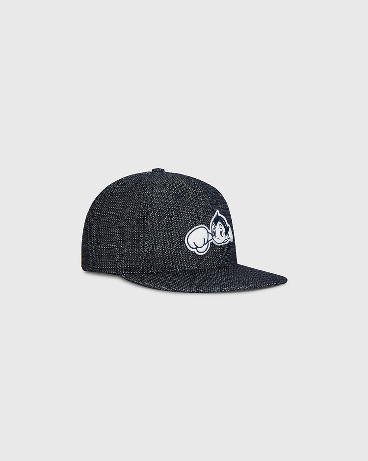 The Sankei Atoms Japanese Indigo Cotton Dobby Ballcap by Ebbets Field features a dark indigo textured fabric and showcases a white embroidered cartoon character peeking over the brim with one arm raised, evoking classic Japanese baseball history. The ballcap is displayed against a plain light gray background.