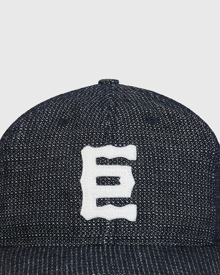 A close-up of the Ebbets Field Tokyo Giants 1940 Japanese Indigo Cotton Ballcap, featuring a dark textured fabric and a white, stylized letter E embroidered on the front. The background is plain and light-colored.