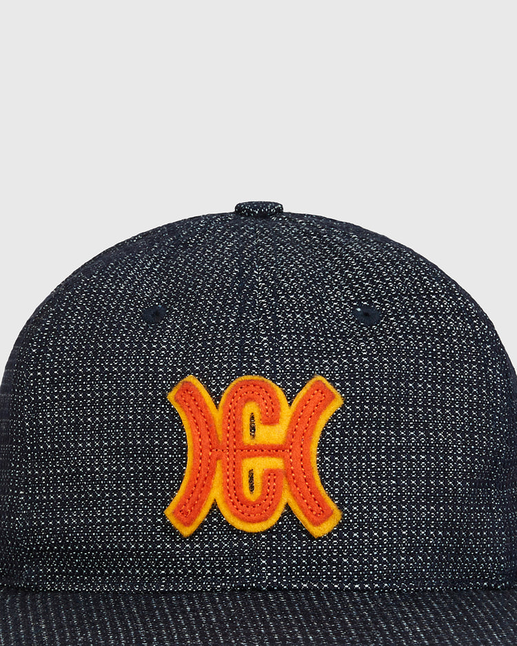 A close-up of the Ebbets Field Hiroshima Carp Japanese Indigo Cotton Dobby Ballcap, crafted from premium Japanese indigo cotton and featuring a striking yellow and red embroidered Hiroshima Carp logo on the front, set against a plain light gray background.