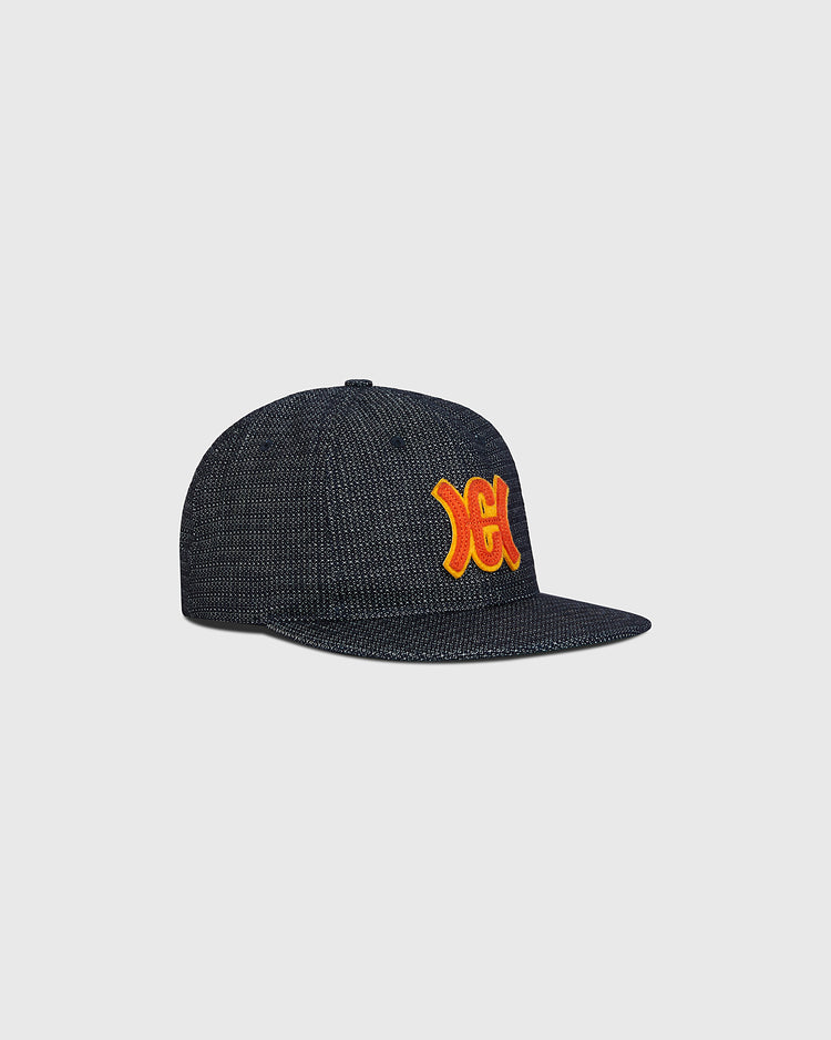 The Hiroshima Carp Japanese Indigo Cotton Dobby Ballcap by Ebbets Field is a dark, textured snapback cap made from premium Japanese cotton, featuring a flat brim and bold yellow and red embroidered “XC” letters on the front. Reminiscent of a classic Hiroshima Carp indigo-dyed ballcap, it is presented against a plain light gray background.