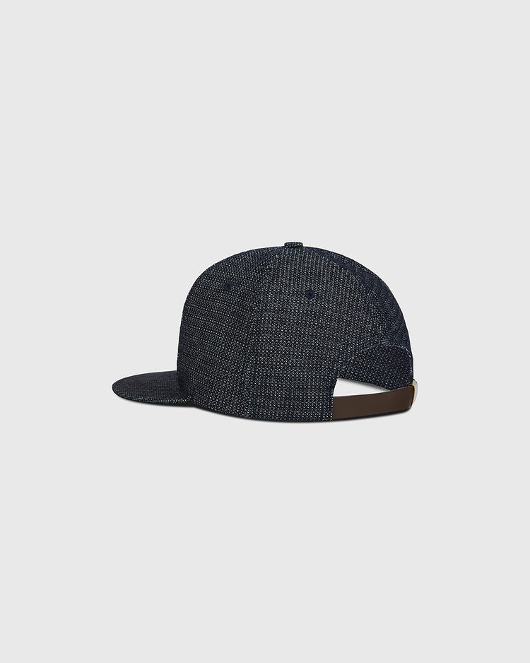 The Ebbets Field Hiroshima Carp Japanese Indigo Cotton Dobby Ballcap is displayed from the back at an angle, showcasing its dark blue indigo-dyed textured Japanese cotton. The ballcap features an adjustable brown strap with a metal clasp, set against a plain light gray background.
