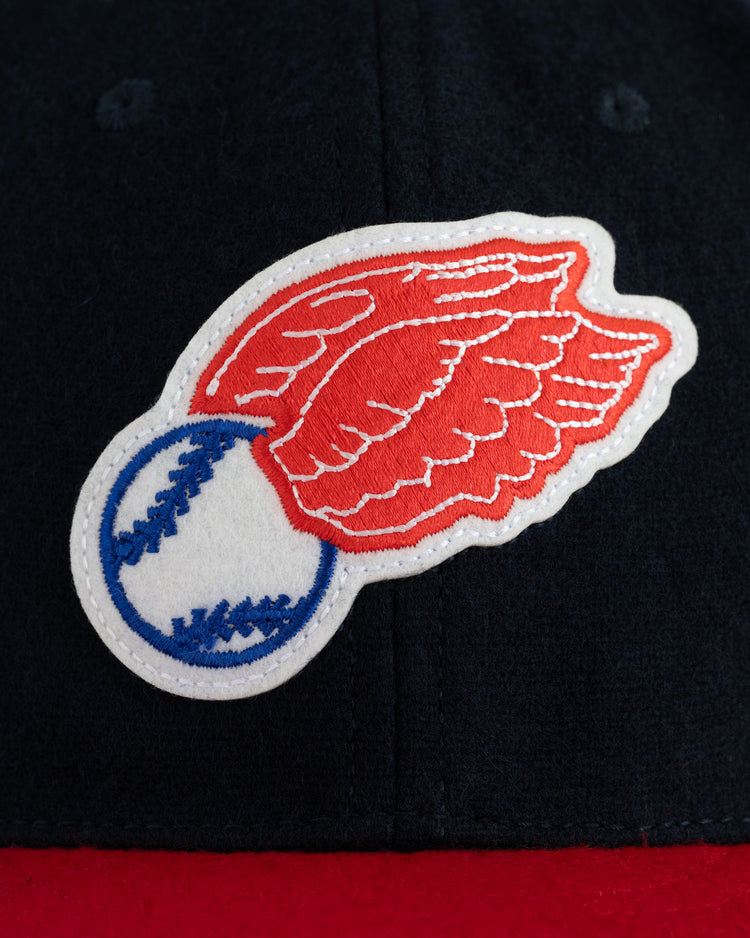 A close-up of the EFF Rochester Red Wings 1950 Authentic Wool Ballcap shows a patch with a white baseball, blue stitching, and red wings sewn onto dark fabric, with a touch of red at the bottom.