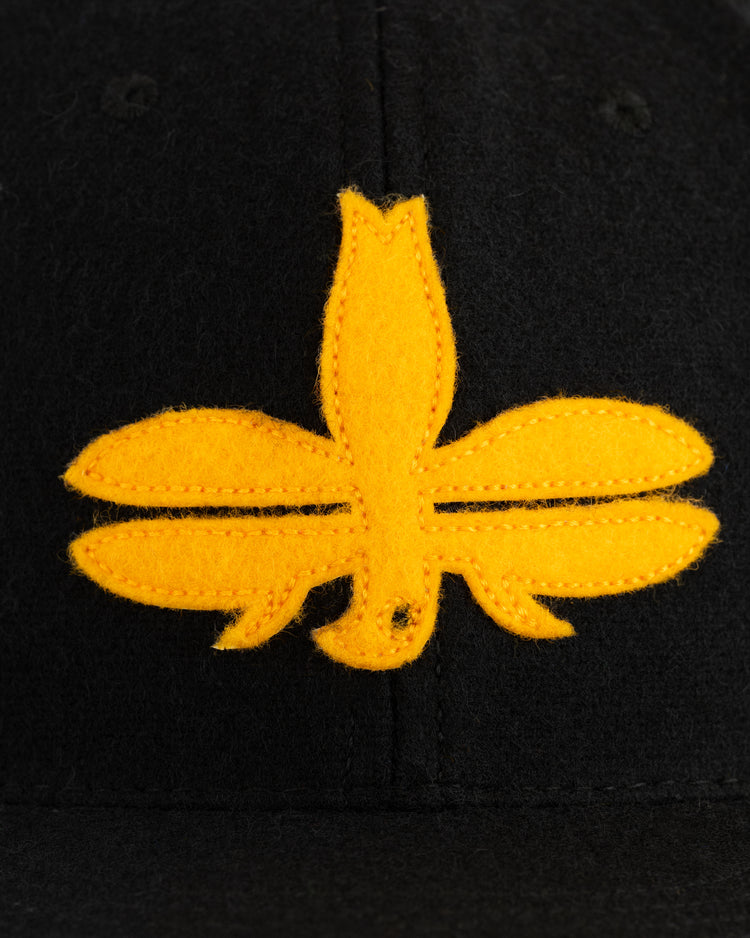 A close-up of a yellow fleur-de-lis patch sewn onto black fabric from the EFF Tombow Unions 1955 Authentic Wool Ballcap, a nod to Japanese baseball history.