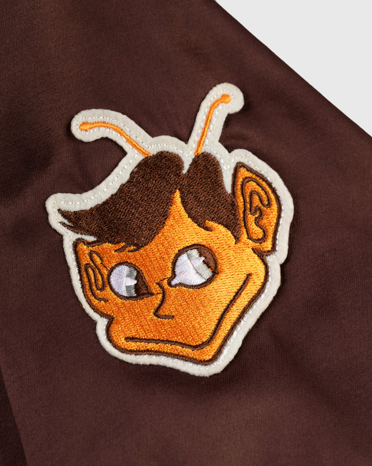 An orange cartoon character patch with short brown hair and antennae, evoking a playful MLB style, is sewn onto a dark brown EFF St. Louis Browns 1952 Satin Windbreaker Jacket.