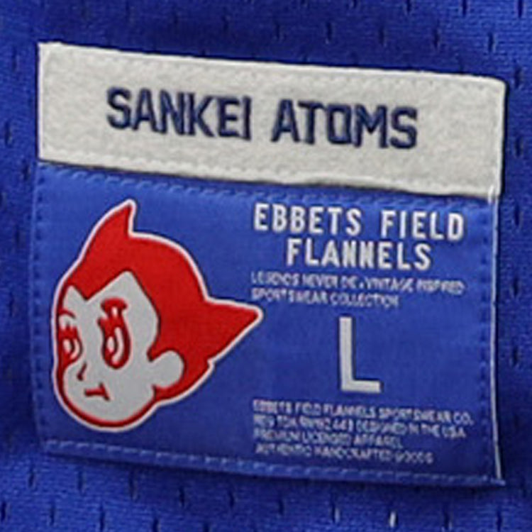 Blue fabric label with SANKEI ATOMS at the top, a cartoon face inspired by Atom Boy, EFF branding below, a large L, and product details referencing the Japanese Central League for the Sankei Atoms EFF DNA Replica V-Neck Mesh Jersey.
