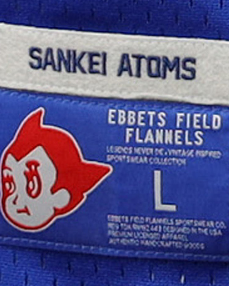 Blue fabric label with SANKEI ATOMS at the top, a cartoon face inspired by Atom Boy, EFF branding below, a large L, and product details referencing the Japanese Central League for the Sankei Atoms EFF DNA Replica V-Neck Mesh Jersey.