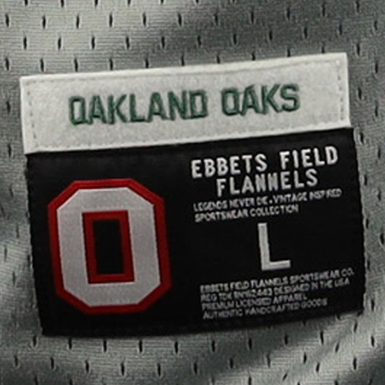 A close-up of the Oakland Oaks EFF DNA Replica Button-Up Mesh Jersey tag shows OAKLAND OAKS in green above a red and white O, honoring their Pacific Coast League legacy, with EFF branding, size L, and product details in white text.