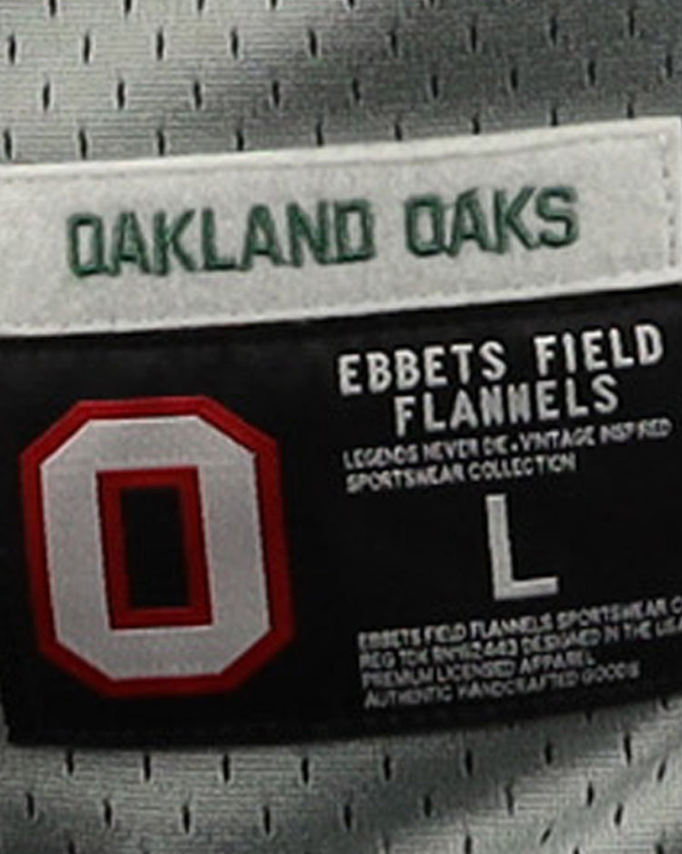 A close-up of the Oakland Oaks EFF DNA Replica Button-Up Mesh Jersey tag shows OAKLAND OAKS in green above a red and white O, honoring their Pacific Coast League legacy, with EFF branding, size L, and product details in white text.