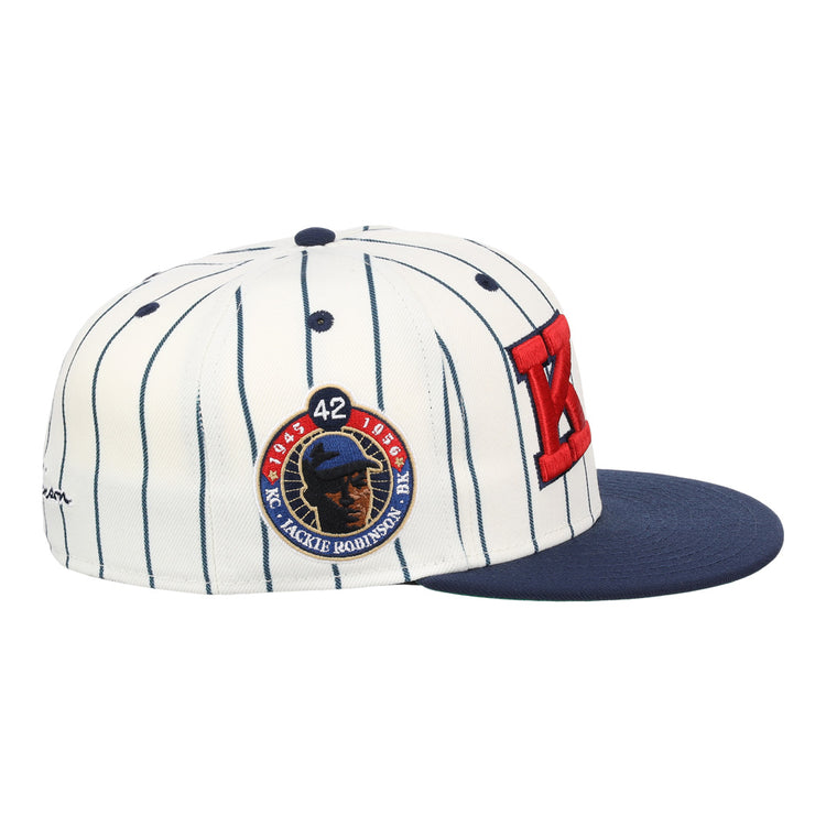 The EFF Kansas City Monarchs Robinson Fitted Ballcap features white fabric with black pinstripes, a navy brim, a bold red K on the side, and a patch showing Jackie Robinson’s face, number 42, and “KC JACKIE ROBINSON.”.