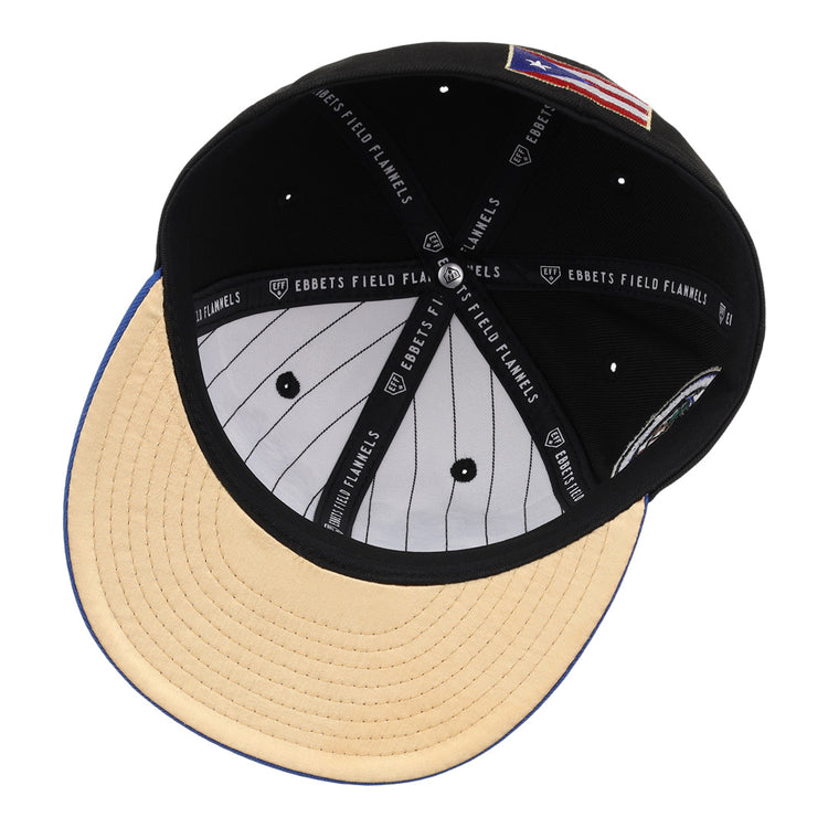 The EFF Santurce Cangrejeros Clemente Fitted Ballcap in black features a beige suede brim, white pinstriped lining, and black sweatband with EBBETS FIELD FLANNELS text. An American flag patch honors Puerto Rican baseball legend Roberto Clemente.