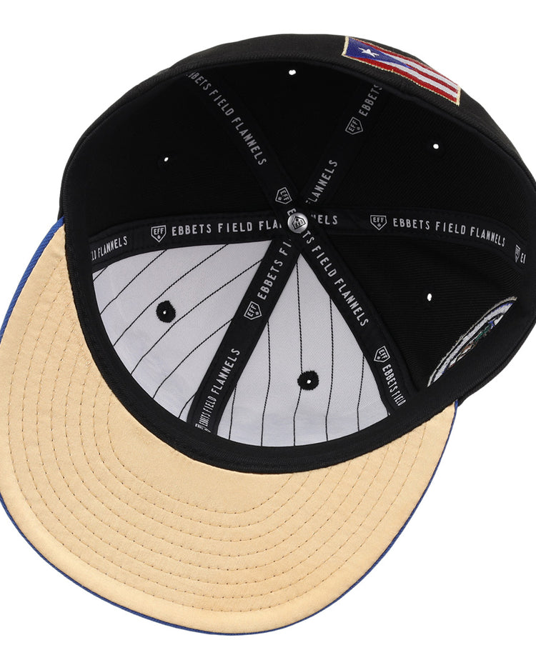 The EFF Santurce Cangrejeros Clemente Fitted Ballcap in black features a beige suede brim, white pinstriped lining, and black sweatband with EBBETS FIELD FLANNELS text. An American flag patch honors Puerto Rican baseball legend Roberto Clemente.