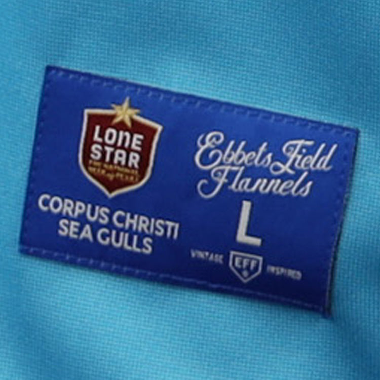 A blue fabric tag on the EFF Corpus Christi Seagulls Lone Star Baseball Jersey features the brand and team name, plus logos in red, white, and gold—celebrating Gulf States League Champions.