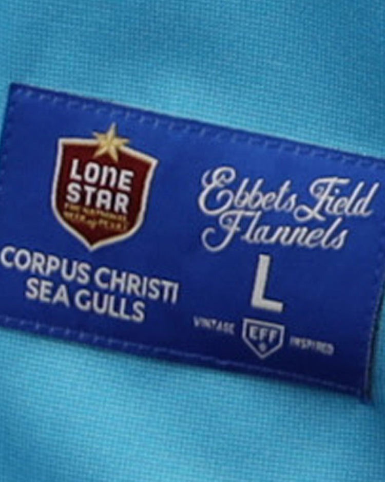 A blue fabric tag on the EFF Corpus Christi Seagulls Lone Star Baseball Jersey features the brand and team name, plus logos in red, white, and gold—celebrating Gulf States League Champions.