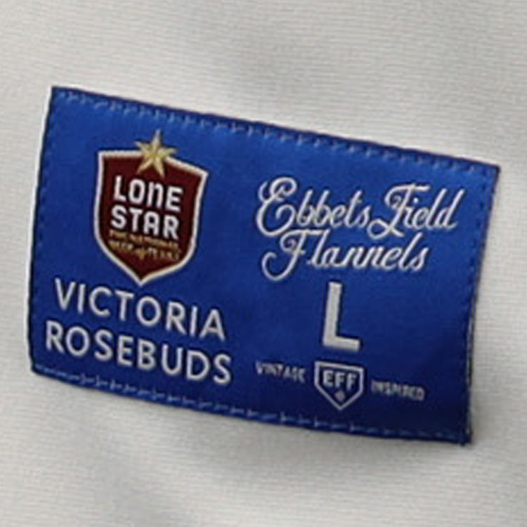 A blue fabric label on the Victoria Rosebuds EFF Lone Star Baseball Jersey by EFF features decorative logos and white text, including Lone Star, Ebbets Field Flannels, Victoria Rosebuds, and L - Vintage Inspired, set on a white garment background.