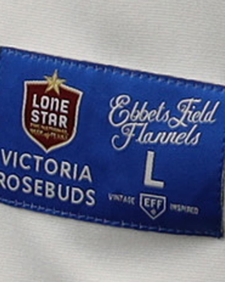 A blue fabric label on the Victoria Rosebuds EFF Lone Star Baseball Jersey by EFF features decorative logos and white text, including Lone Star, Ebbets Field Flannels, Victoria Rosebuds, and L - Vintage Inspired, set on a white garment background.