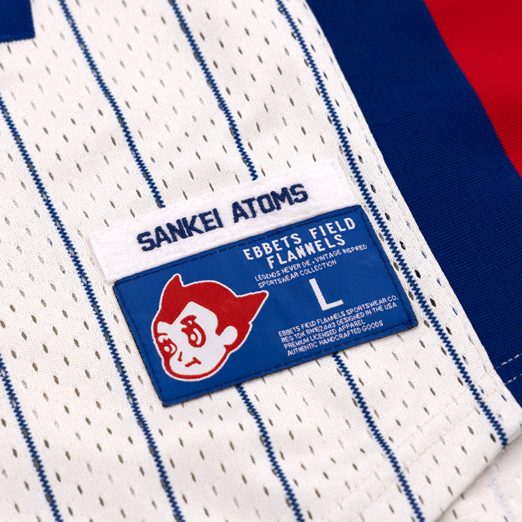 A close-up of the EFF Sankei Atoms Pinstripe Button Down Jersey shows a blue Sankei Atoms tag and an Ebbets Field Flannels patch with a cartoon character, with red and blue fabric in the background.