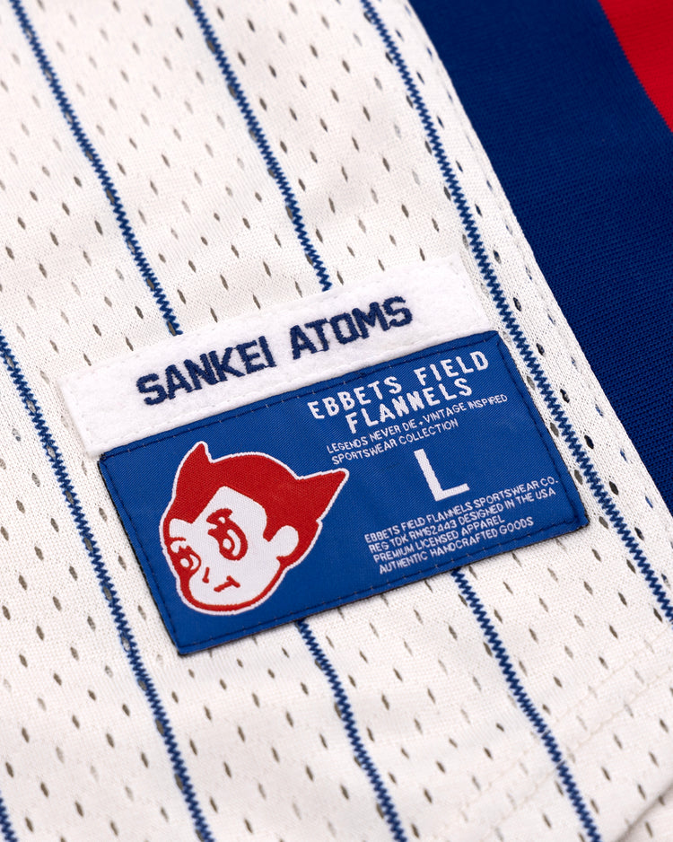 A close-up of the EFF Sankei Atoms Pinstripe Button Down Jersey shows a blue Sankei Atoms tag and an Ebbets Field Flannels patch with a cartoon character, with red and blue fabric in the background.