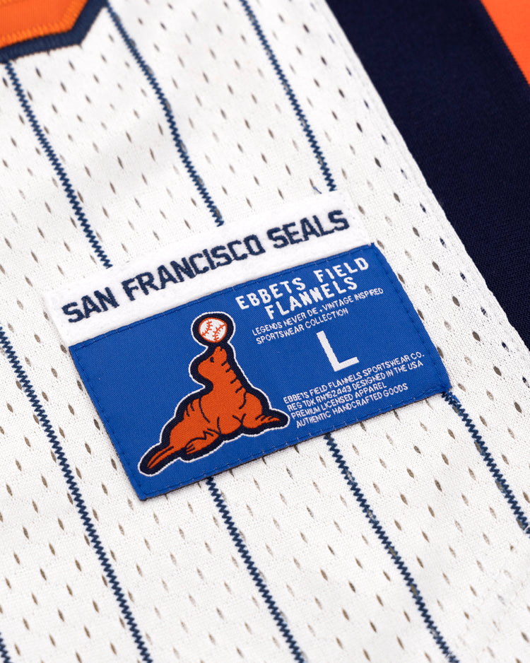 A close-up of the EFF San Francisco Seals Pinstripe Button Down Jersey label, showing a blue tag with orange seal logo, size L, and EFF branding on a white pinstripe jersey with blue accents and navy/orange trim.