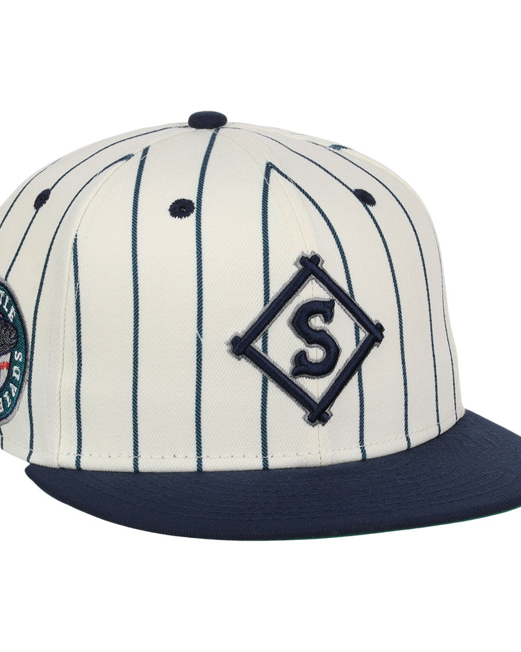 The EFF Seattle Steelheads NLB Pinstripe Fitted Ballcap features a cream base with navy pinstripes, a navy bill, an embroidered S in a diamond on the front, and a Seattle Mariners patch on the side.