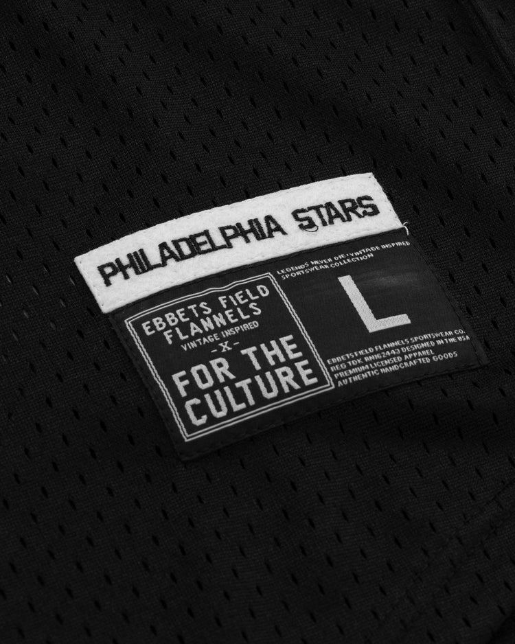A close-up of black mesh fabric with two labels: one celebrates the Philadelphia Stars and EFF’s Cultural Excellence x Philadelphia Stars Button Down Jersey, while the other highlights Ebbets Field Flannels, For the Culture, and size L.