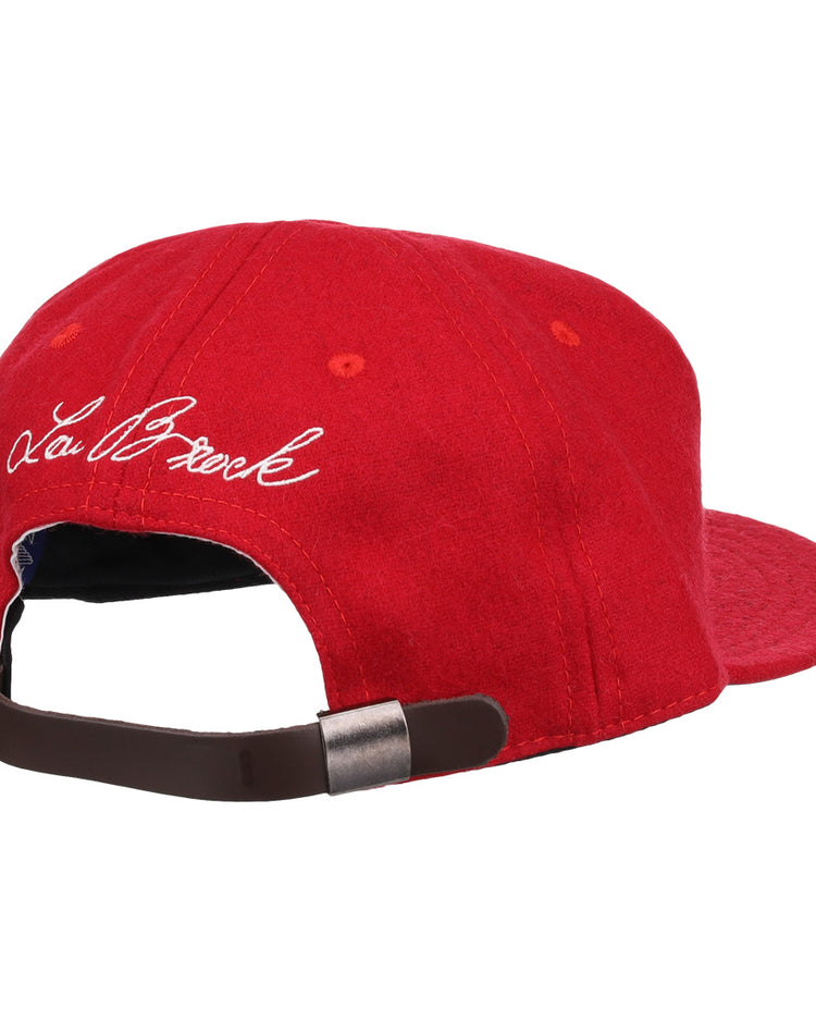 The Ebbets Field Flannels Lou Brock Signature Series Ballcap is a red vintage wool cap with a brown adjustable strap and metal clasp. White cursive embroidery of the Lou Brock signature appears on the left side.